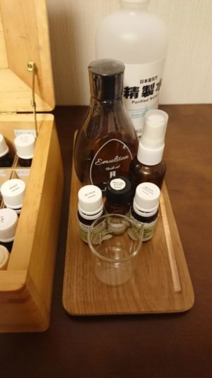 精製水、乳化剤、精油、ビーカーなど、防腐剤なしのアロマ化粧水を作るための材料が木製トレーに並んでいる様子。