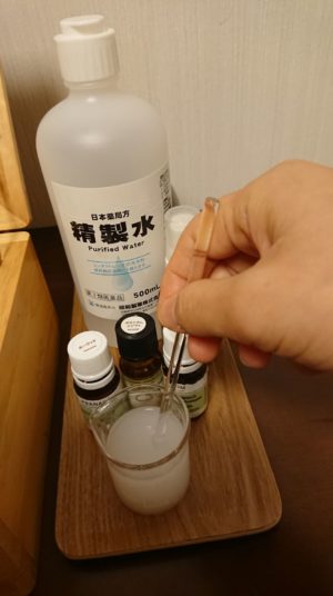 ビーカーの中の乳白色の化粧水を、ガラス棒で手が静かに混ぜているアロマ化粧水の製作工程。