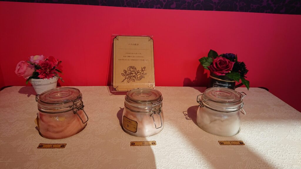 におい展のローズ3種の嗅ぎ比べ展示。A・B・Cのラベルが付いたガラス瓶がテーブルに並び、それぞれの香りを体験できる様子。