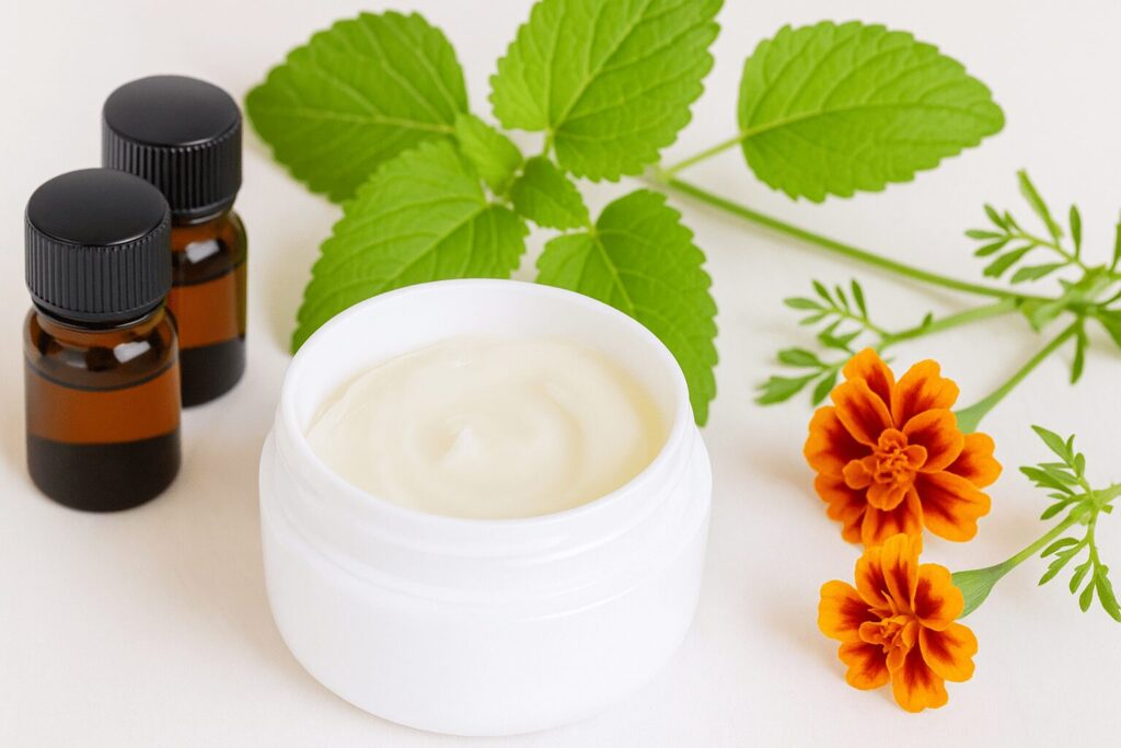 白い容器のアロマジェルと遮光瓶、レモンバーベナの葉とオレンジ色の花を並べた写真
