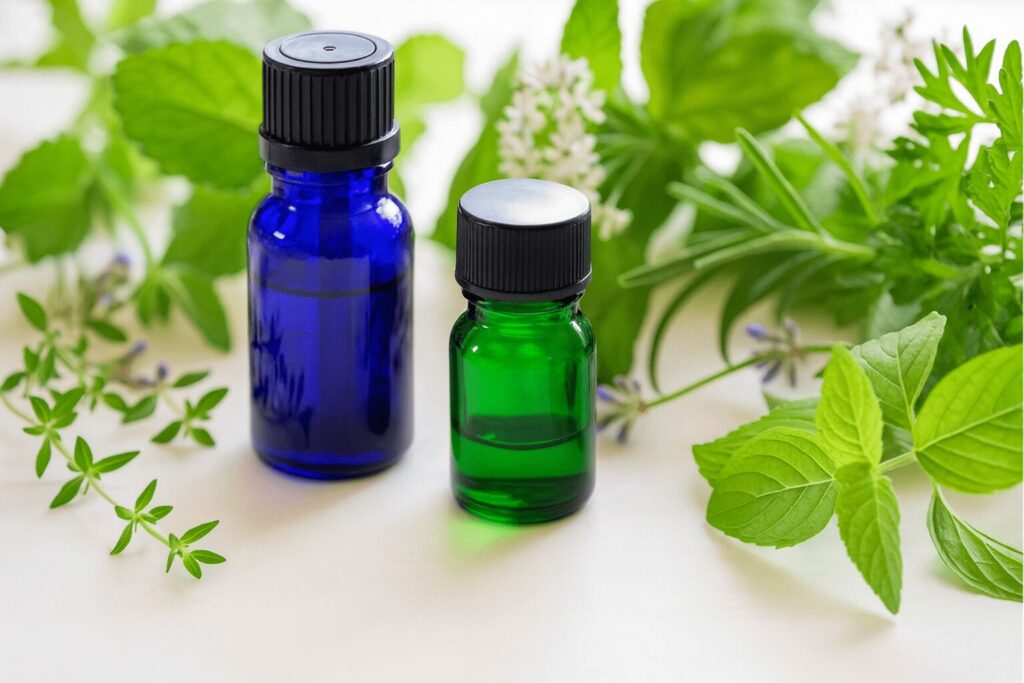 青と緑の遮光瓶とハーブが白いテーブルに並んだ精油のイメージ写真