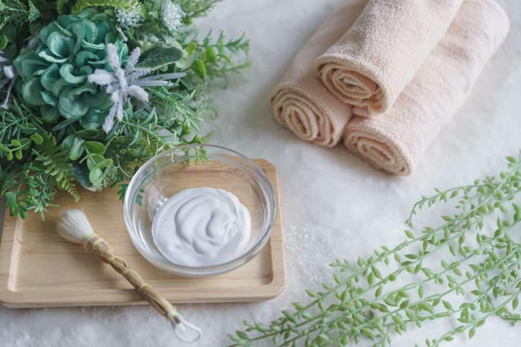 フェイシャル用のクリームと筆、タオル、グリーンの葉を並べたエステイメージ写真。肌悩みに合わせたフェイシャルケアを表現。