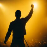 スポットライトを浴びながら観客に腕を伸ばすステージ上の男性シルエットの写真。フレディ・マーキュリーを想起させる力強いポーズ。